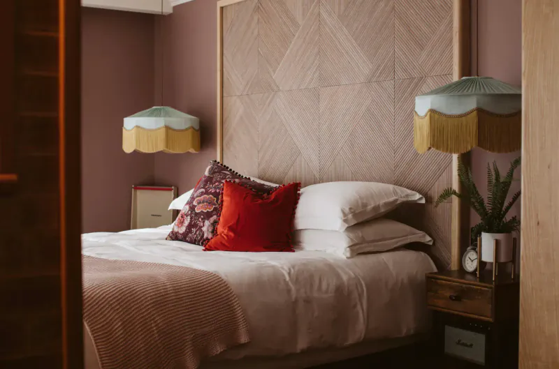 Cozy bedroom at The Rabbit Hotel with wooden headboard, colorful pillows, white bedding, fringed lamps, and plants.