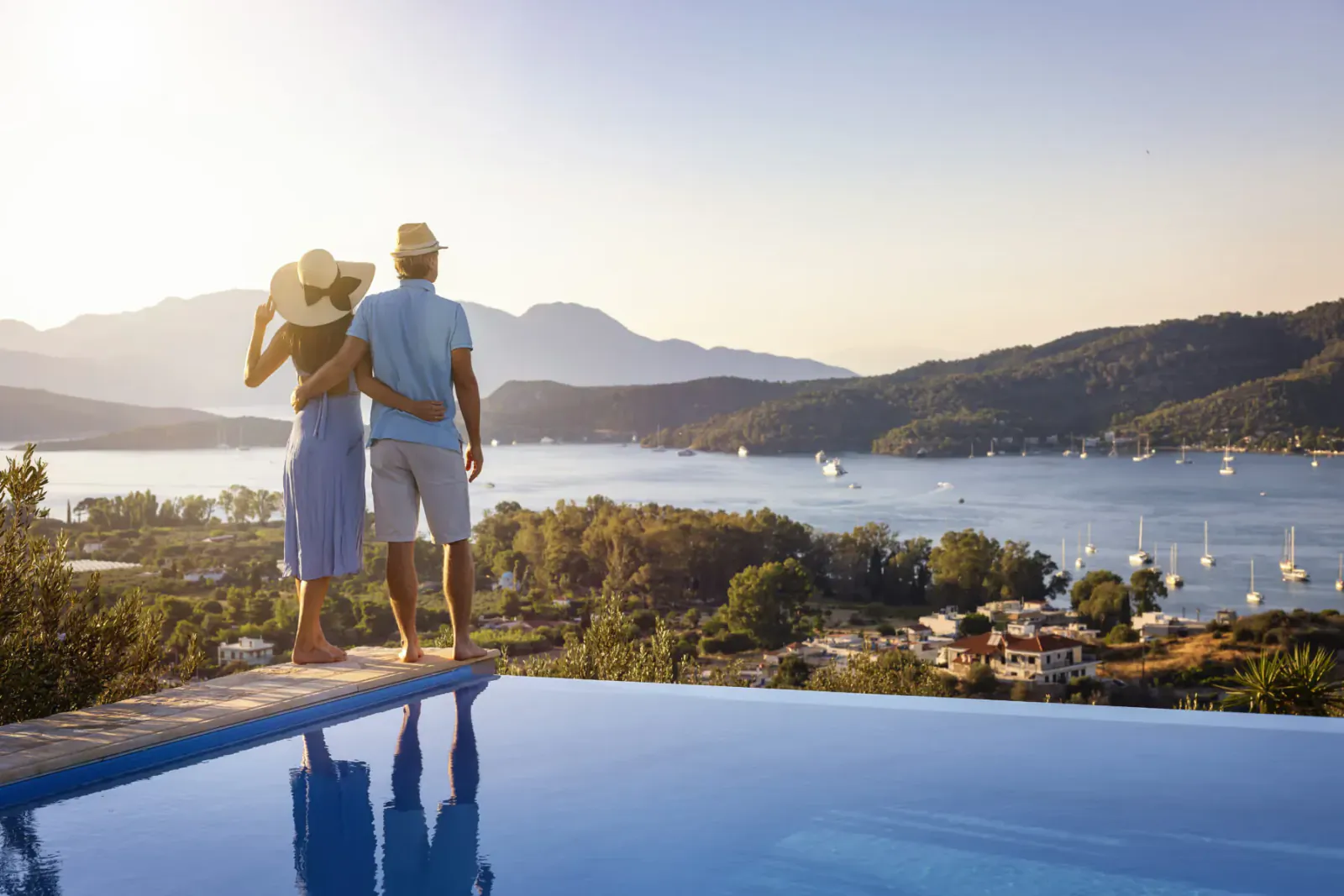 Couple in sun hats and summer clothes stand arm-in-arm at infinity pool edge, overlooking lake, sailboats, mountains at sunset.