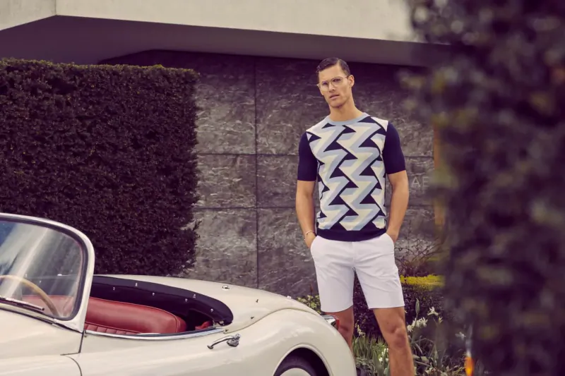 Man in zigzag short-sleeve shirt and white shorts stands by white convertible sports car against stone wall and hedges.
