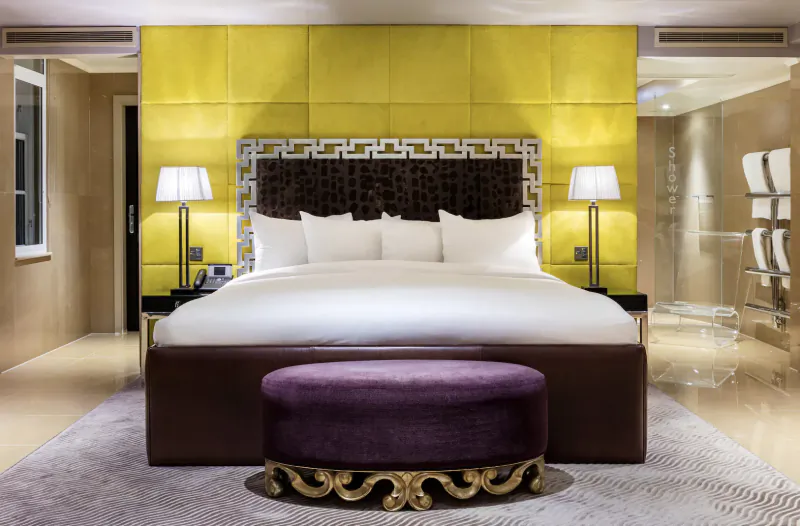 Luxurious hotel bedroom with king bed, yellow quilted wall, lamps, purple ottoman, and marble floors at The May Fair Hotel.