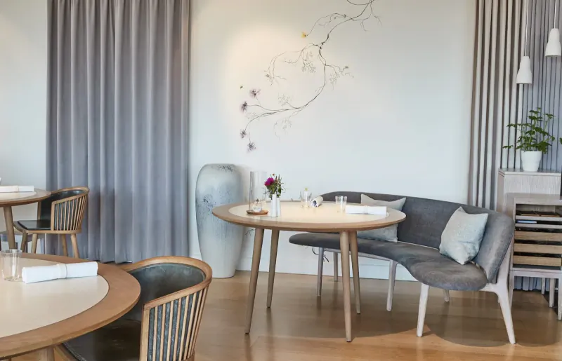 Geranium Restaurant interior: modern dining area with round wooden table set for meal, curved sofa, floral wall art, plants, gray curtains.