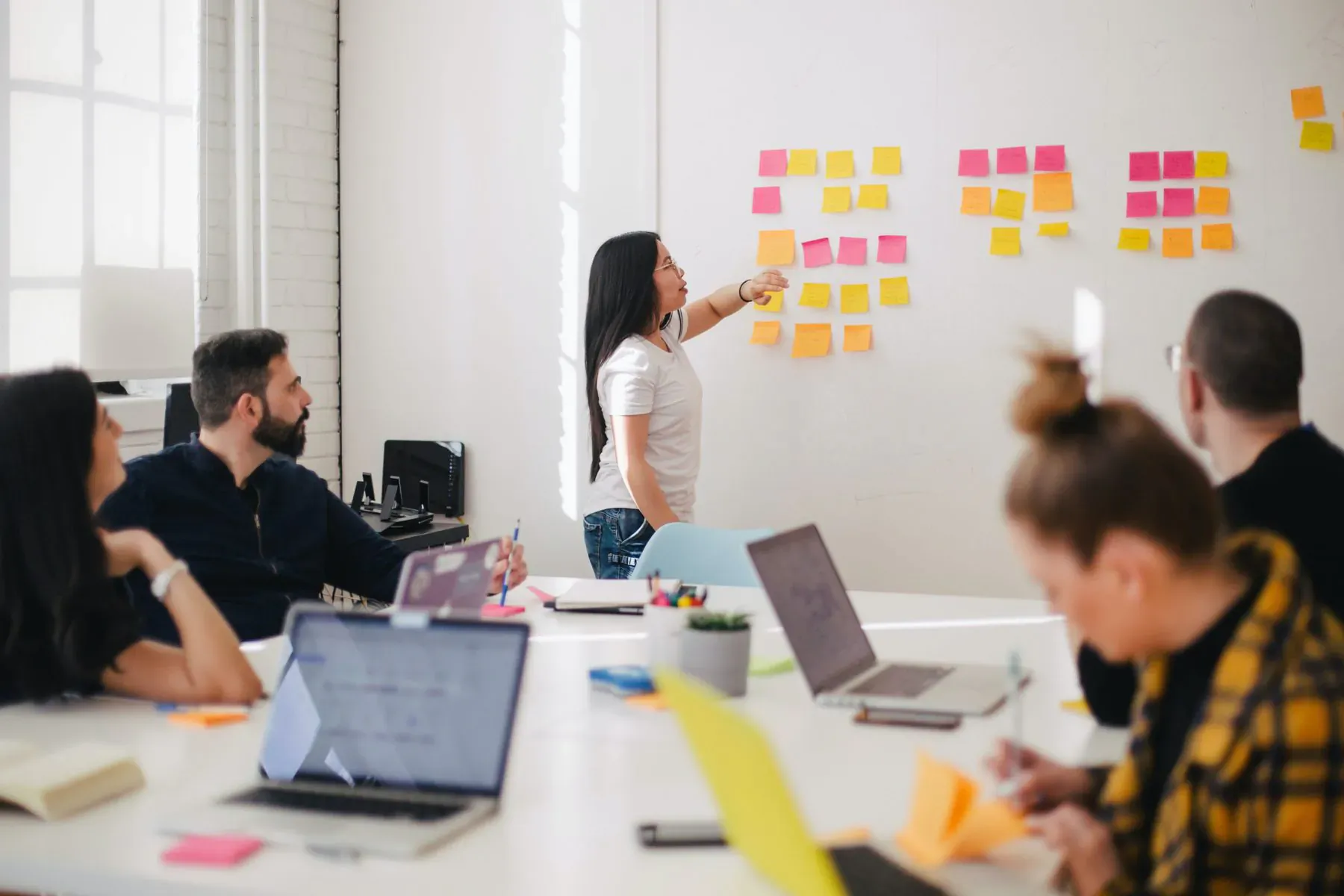 Woman presenting yellow sticky notes on whiteboard to diverse team at conference table with laptops