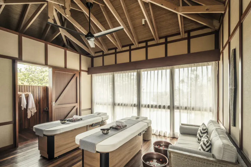 Spacious wooden massage room in Four Seasons Desroches villa with two treatment tables, towels, basins, and garden view.