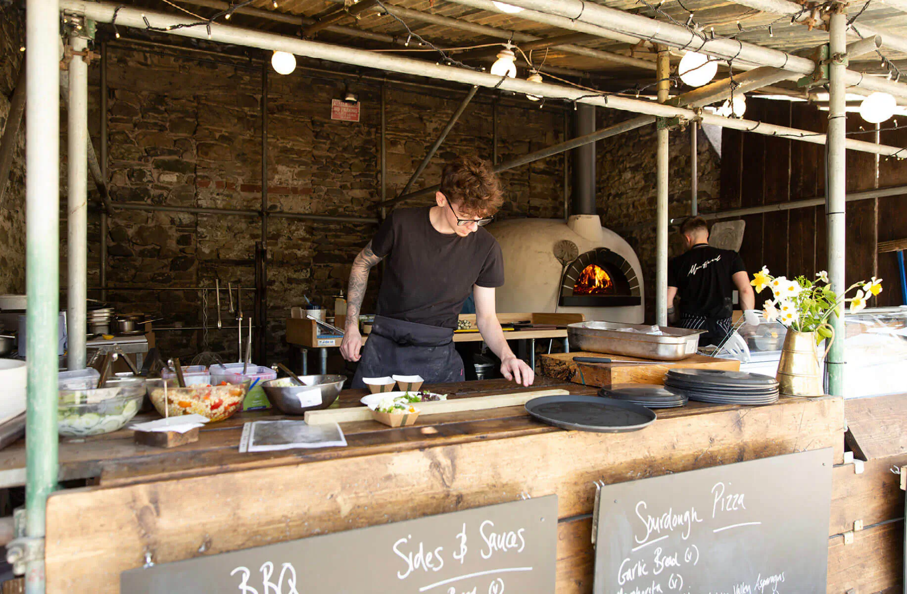 Tattooed chef with glasses works at outdoor pizza stall with wood-fired oven; chalkboard reads 'Saus & Saus Pizza £8.50'