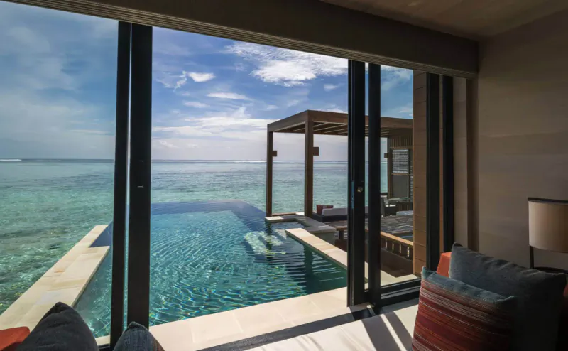Luxury overwater villa interior with sliding doors open to turquoise lagoon and infinity pool, Maldives.