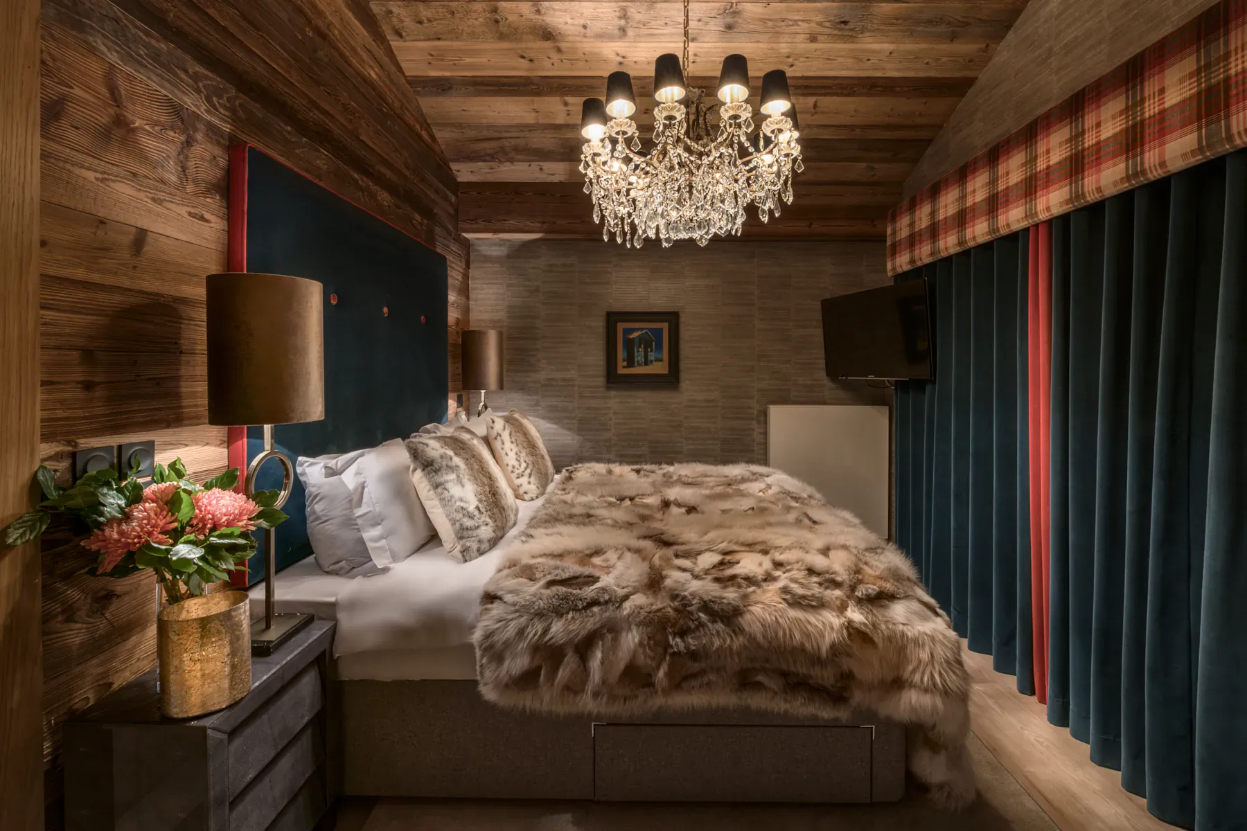 Luxurious mountainside chalet bedroom with fur-covered bed, chandelier, plaid curtains, and flowers.