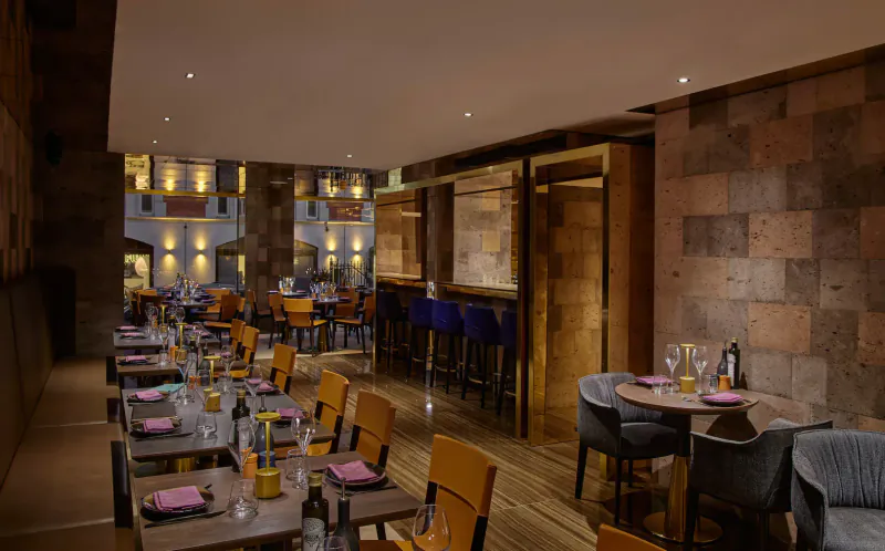 Elegant restaurant interior at Lusin Mayfair with wooden tables set with pink napkins, orange chairs, blue bar stools, and stone walls.