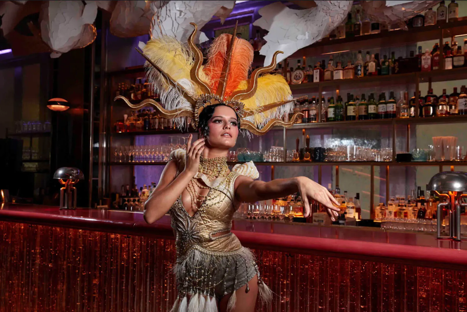Woman in ornate feathered headdress and gold fringe dress dances at illuminated bar with liquor shelves