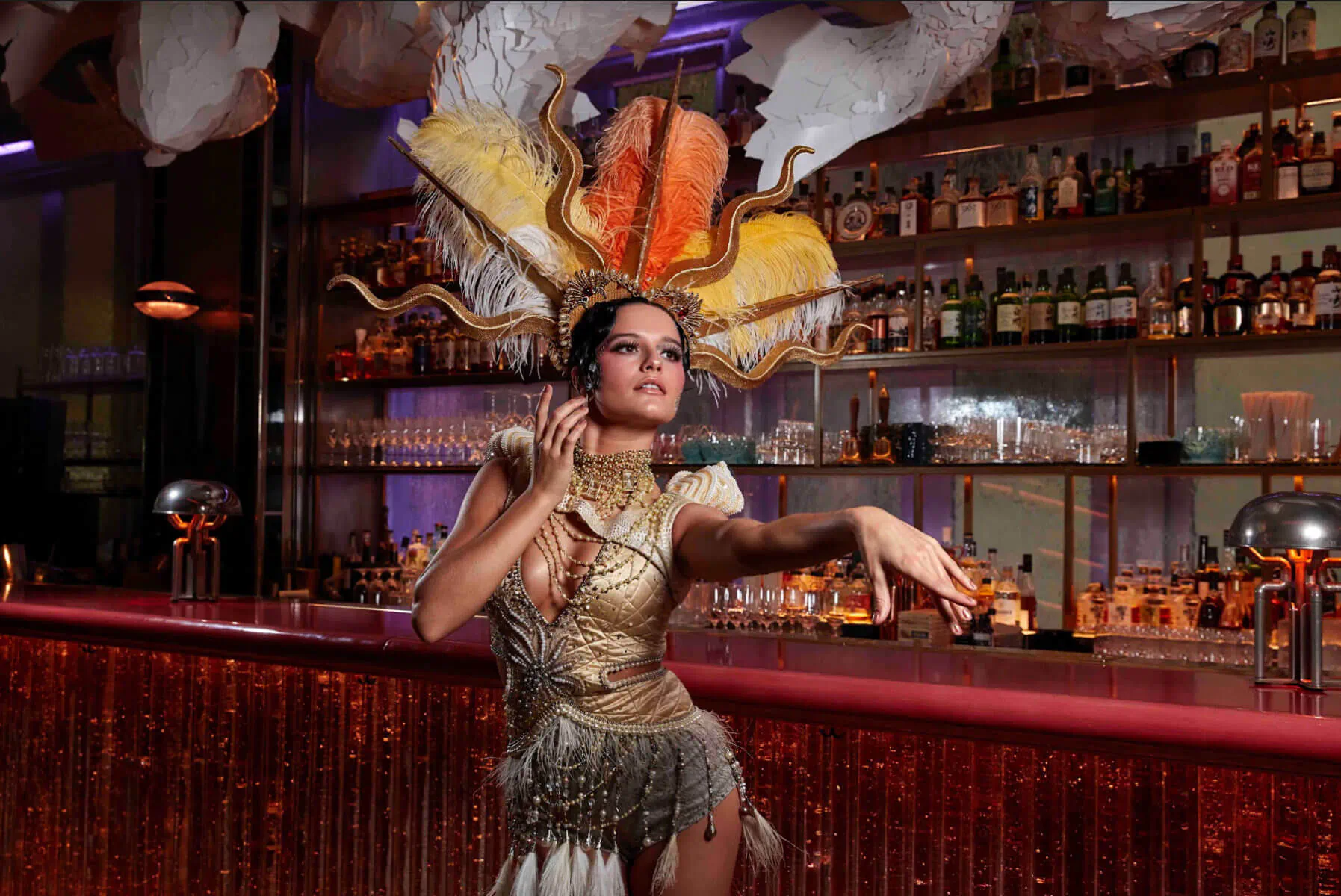Woman in ornate feathered headdress and gold fringe dress dances at illuminated bar with liquor shelves
