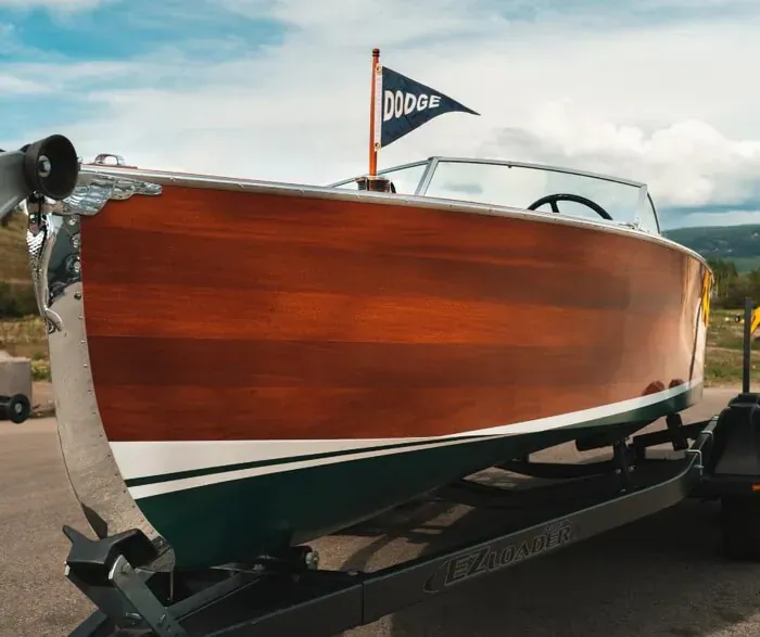 Polished wooden Dodge speedboat on trailer with Dodge flag, under blue sky, in need of replacement.