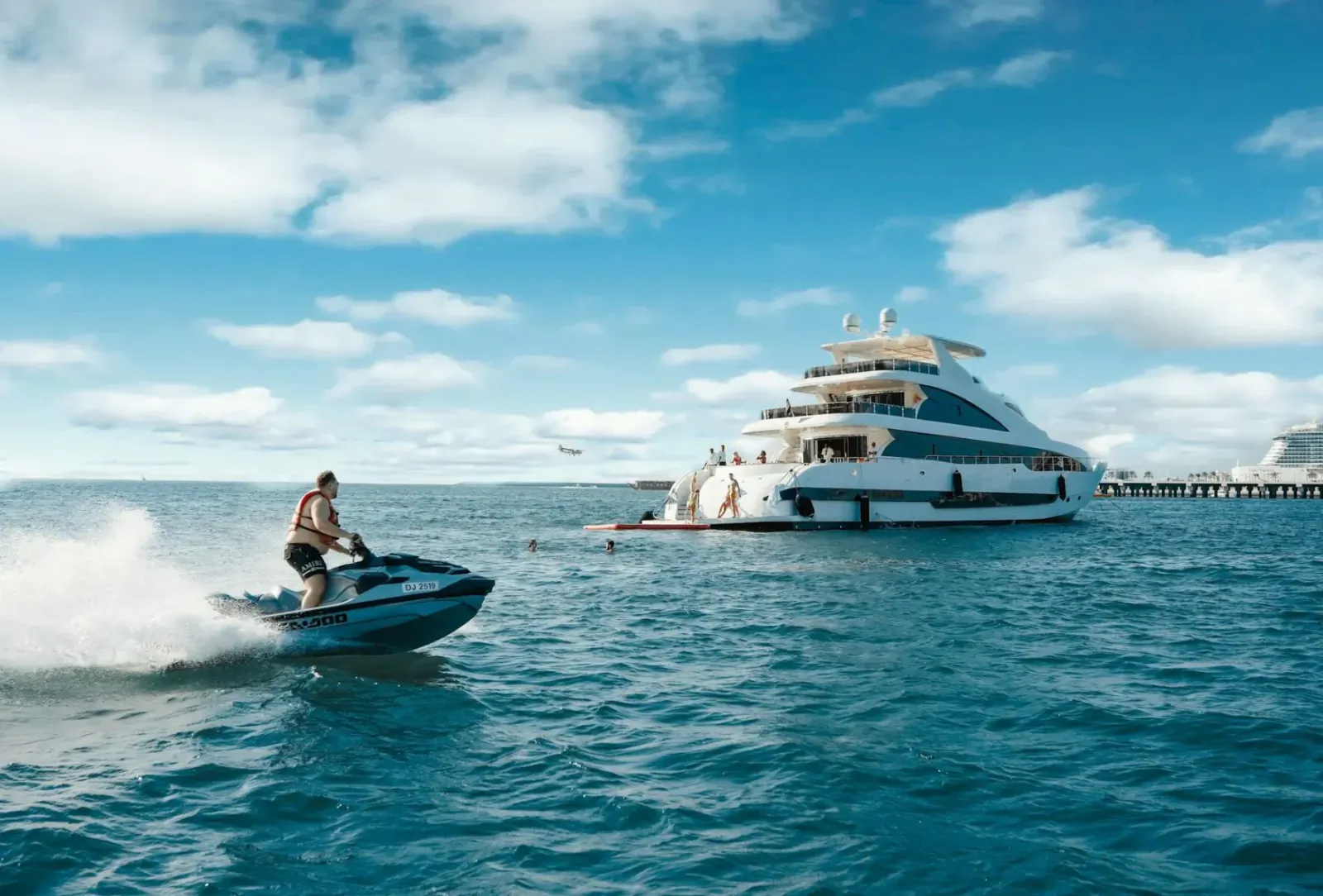 Man riding jet ski with water spray near large white luxury yacht on blue ocean under partly cloudy sky