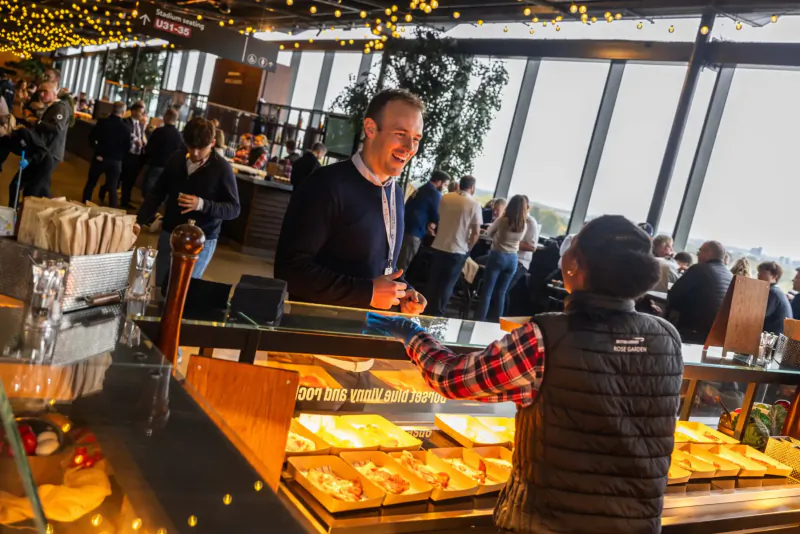 Man in badge smiling as server in black vest hands him pastry from buffet counter in festive rooftop gallery with crowd.