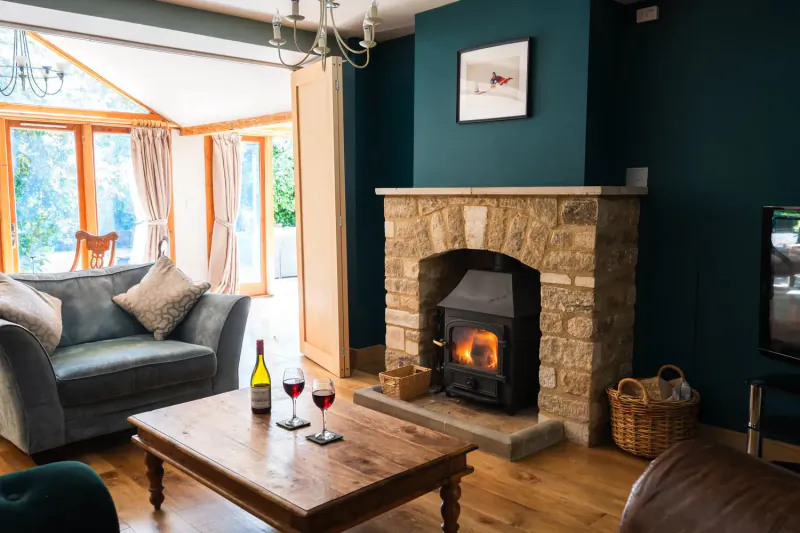 Cozy Cotswolds living room with lit stone fireplace, teal walls, gray sofa, coffee table with wine glasses, garden view.