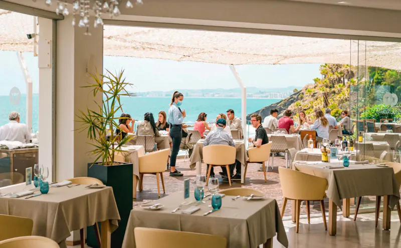 Busy seaside restaurant with diners at tables, server attending, ocean view through large windows and terrace.
