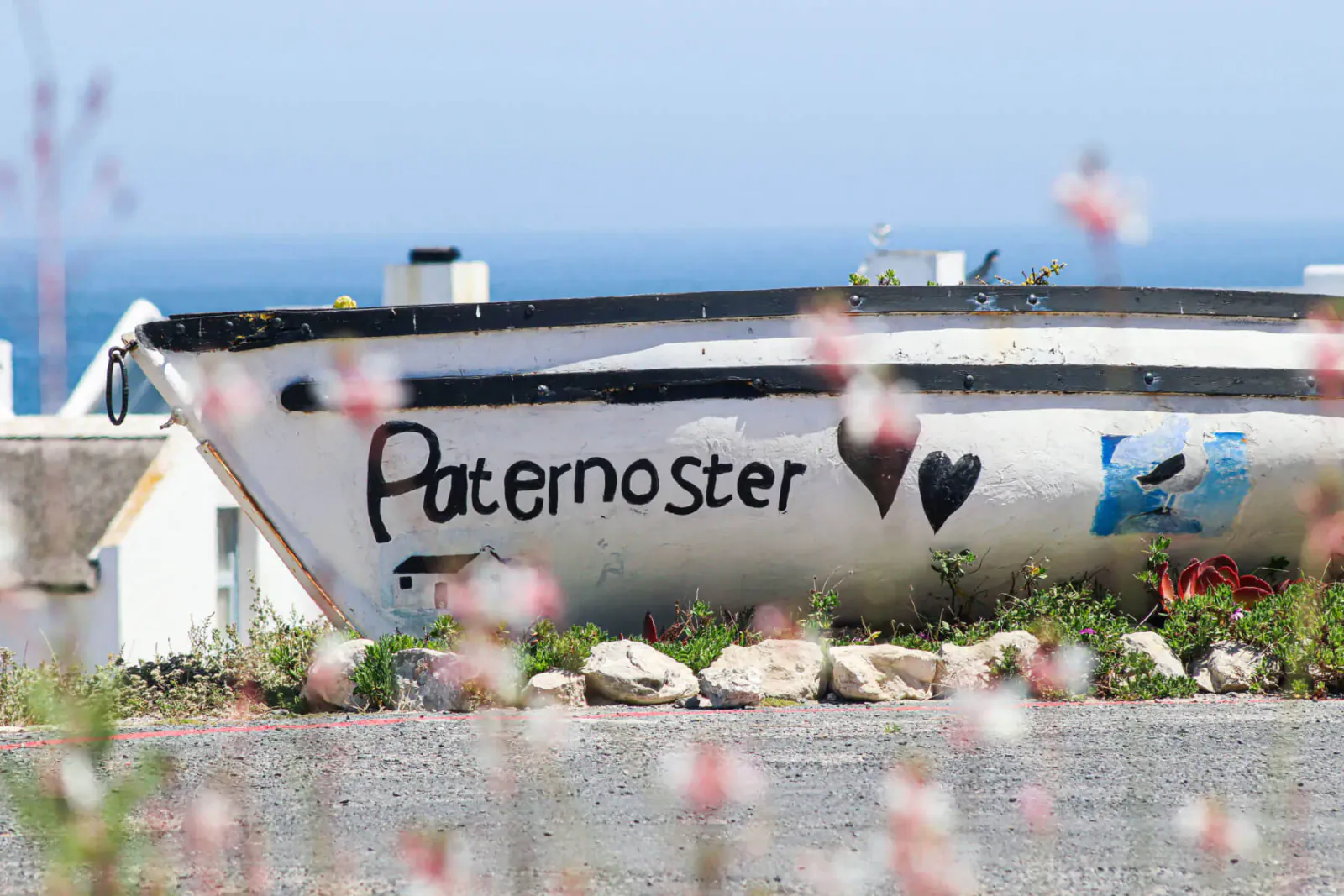 White fishing boat named 'Paternoster' with heart and blue emblem, on shore by ocean, flowers in foreground.