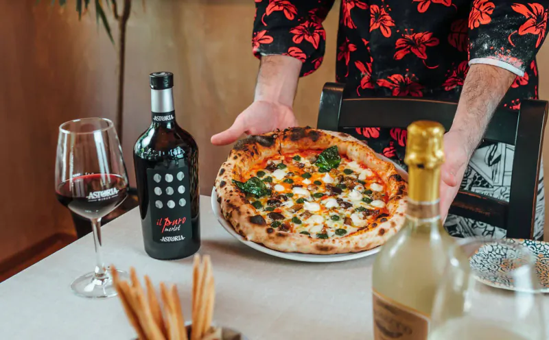 Person in red floral shirt holding 'La Pala D’Oro' pizza with burrata and basil, beside wine glass, Amarone bottle, and sparkling wine on table.