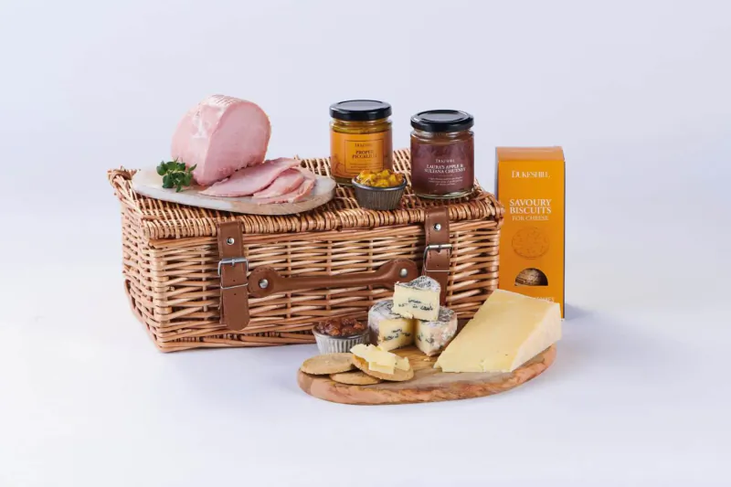 Open wicker picnic hamper with sliced ham, cheeses, chutneys, crackers, and Tiptree jar on white background.