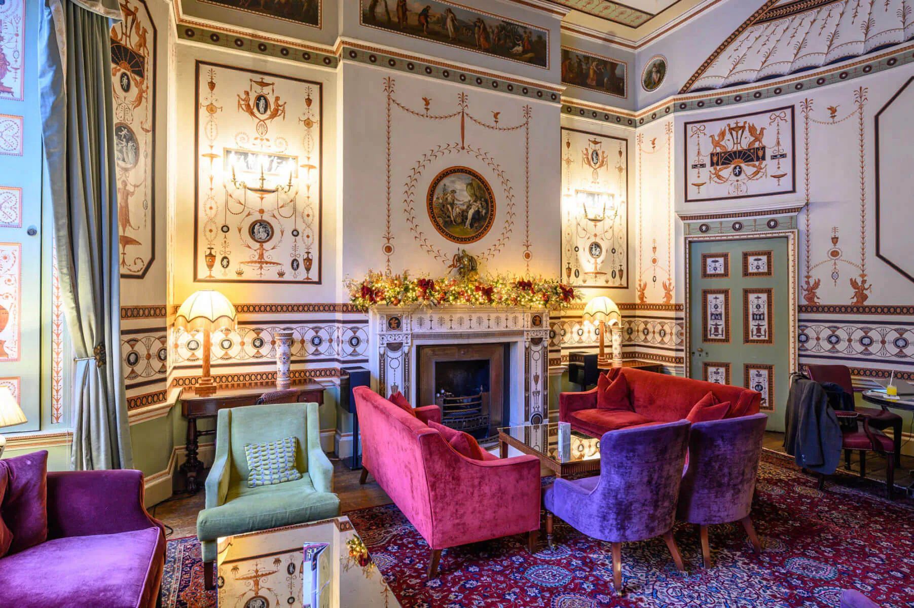 Festive drawing room at Home House, London: ornate walls, pink velvet sofas, green chairs, lit fireplace, garland.