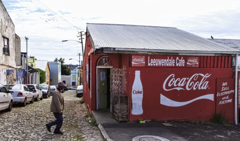 Man walking past Leewendale Cafe with large Coca-Cola sign on colorful Bo-Kaap street.