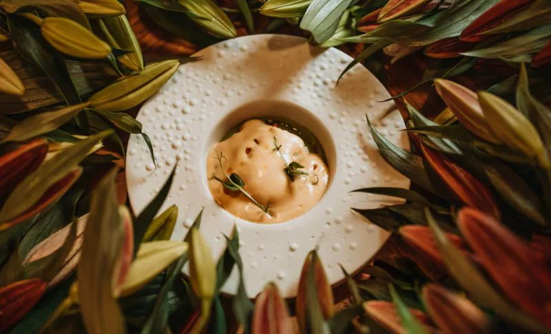 Creamy dish with herbs in white bowl, surrounded by green and orange lily flowers on plate