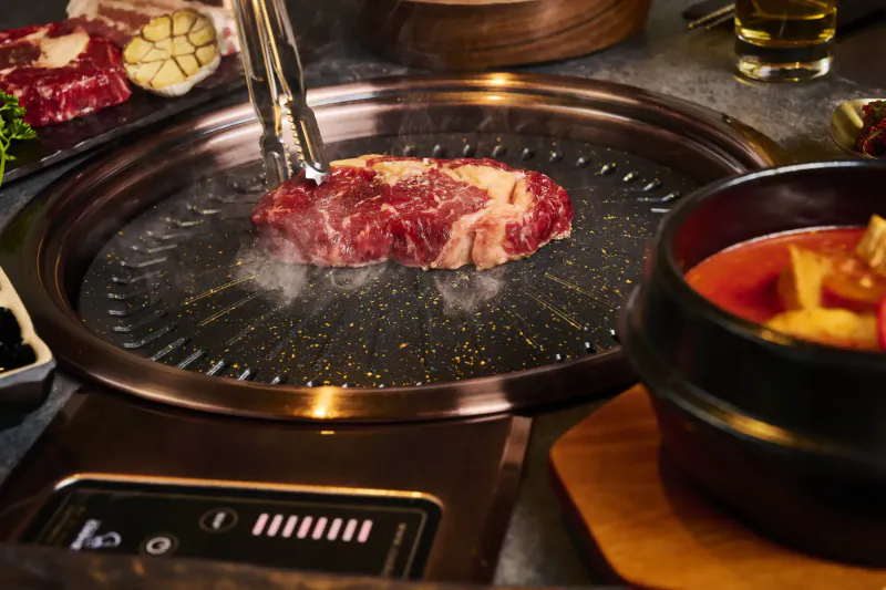 Sizzling marbled steak on Korean BBQ grill with tongs, garnishes, garlic, and red soup bowl nearby.