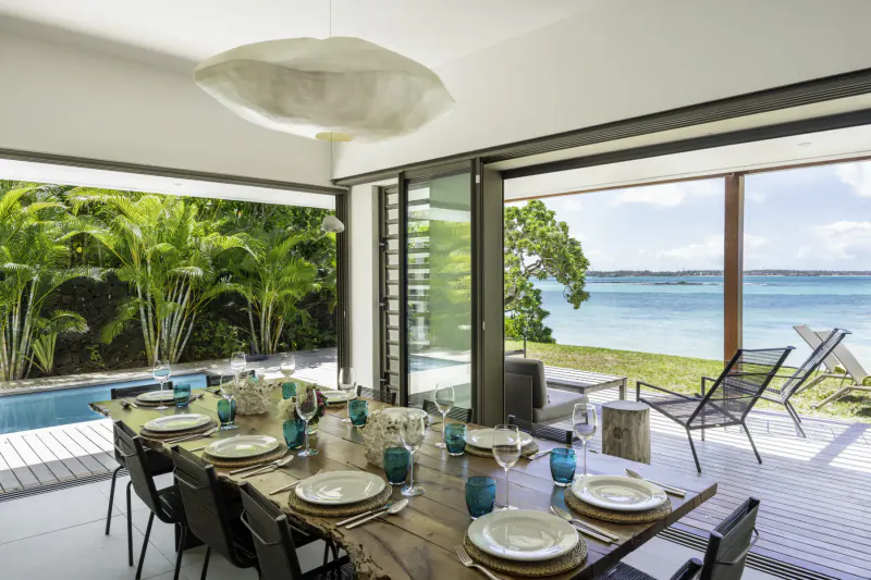 Elegant dining table set for meal in modern villa, open doors to lush garden, infinity pool, and turquoise Belle Mare beach.