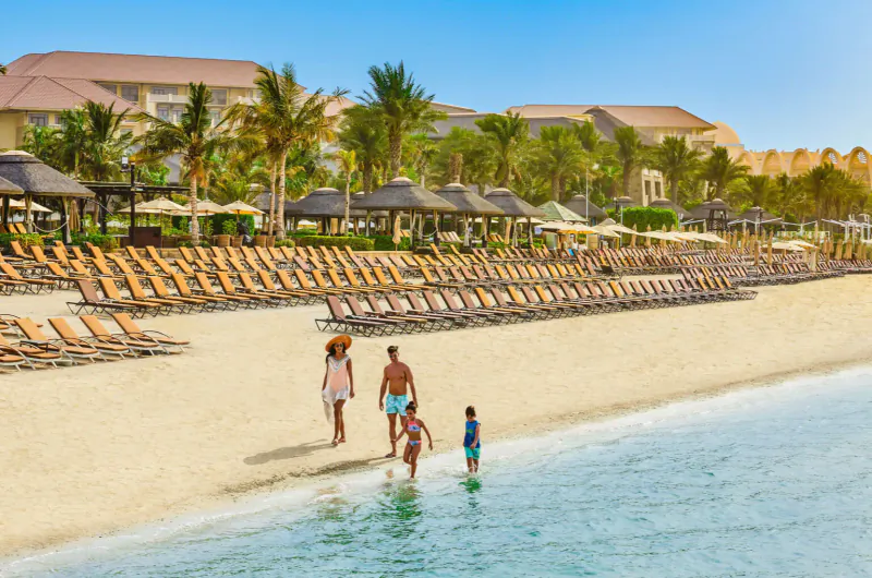 Family walking on Sofitel Dubai The Palm beach with lounge chairs, palm trees, and resort buildings by the sea.