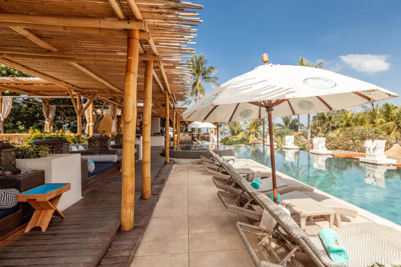Azul Beach Club Bali: tropical infinity pool with loungers, white umbrellas, bamboo lounge under blue sky and palms.