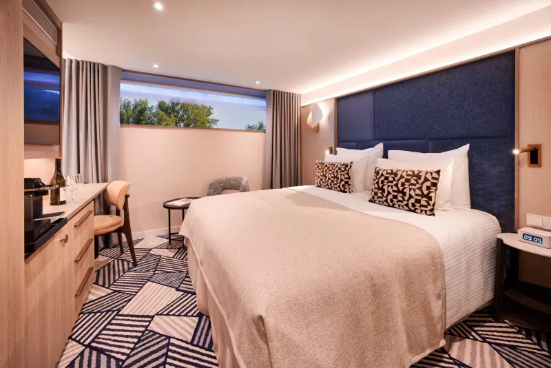 Elegant river cruise stateroom with king bed, leopard pillows, wood desk, window at dusk overlooking Netherlands.