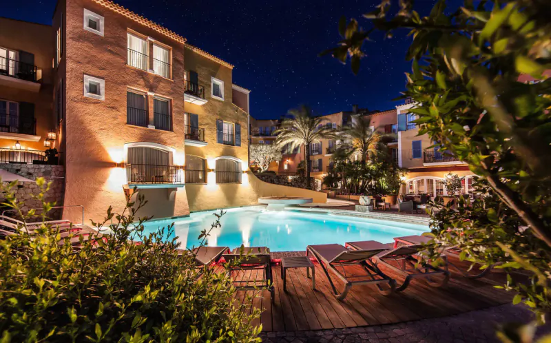 Nighttime view of Byblos Palace hotel pool in Saint-Tropez, with lit terracotta buildings, palm trees, and lounge chairs.