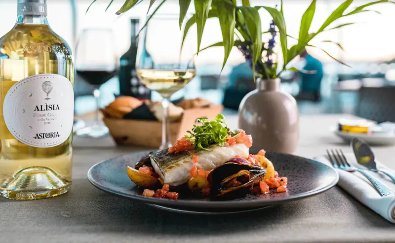 Sea bass and mussels on black plate with tomato sauce, Alenia white wine bottle, glasses, seaside terrace view
