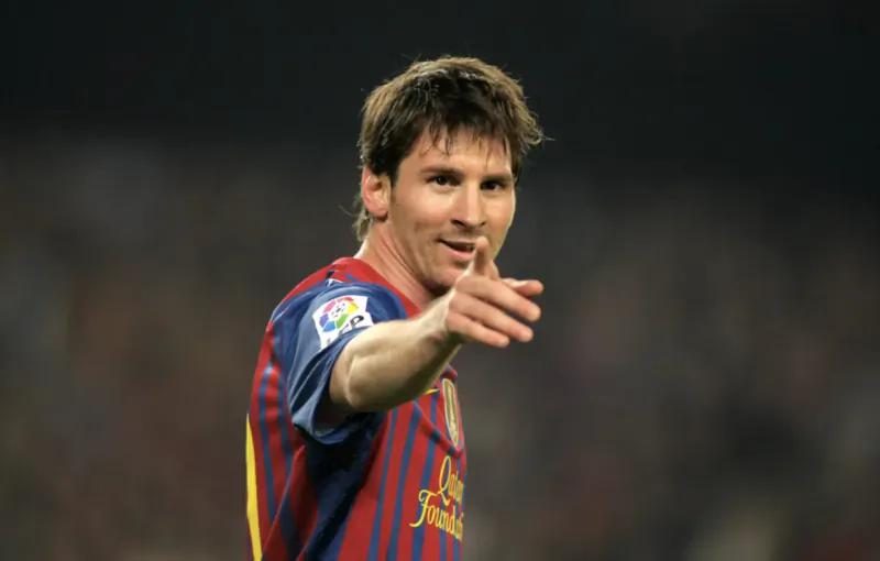 Lionel Messi in FC Barcelona jersey, pointing on soccer field at night stadium.