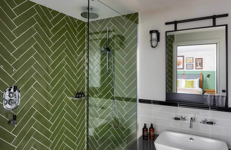 Contemporary hotel bathroom with green herringbone tiled walk-in shower, white vanity sink, mirror, and bedside lamp reflection.