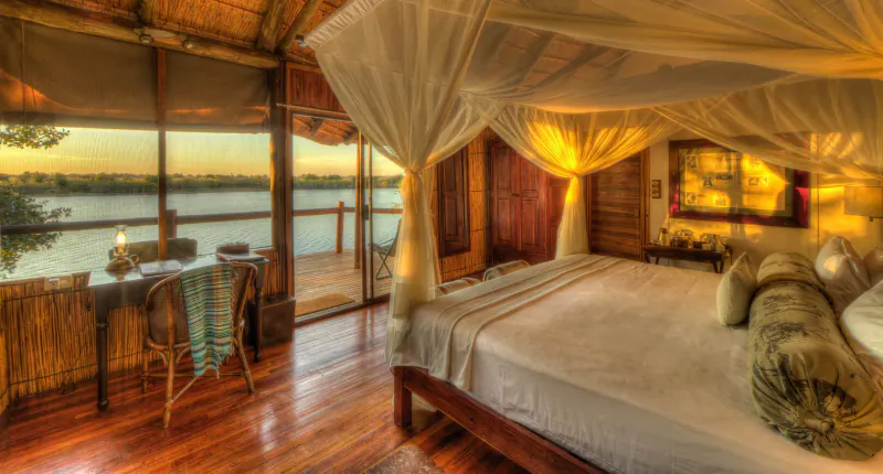 Luxurious bedroom in Xugana Island Lodge with mosquito-net canopy bed, wooden deck overlooking serene lake at sunset.