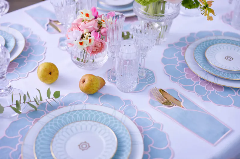 Luxury table setting with blue floral placemats, ornate plates, pink roses, pears, and glassware on white tablecloth