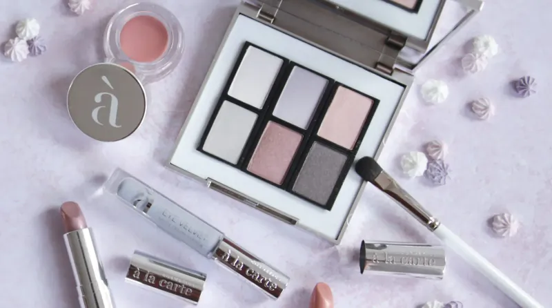 Open À la Carte eyeshadow palette with gray shades, lipstick, lip gloss, brush on pink surface with candy sprinkles