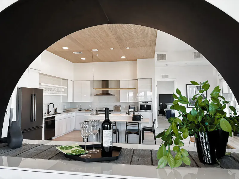 Modern white kitchen in Ariva Residences with wood ceiling, viewed through black arch; counter holds wine glasses, bottle, charcuterie, plants.