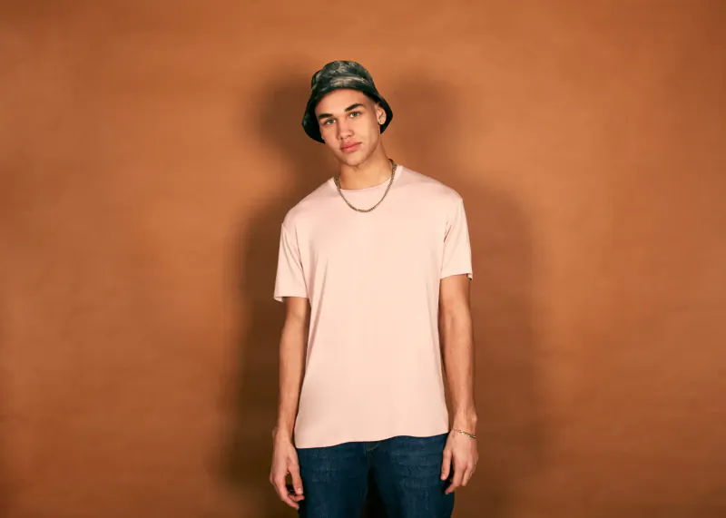 Young man with tan skin wearing camouflage bucket hat, white t-shirt, silver chain, bracelet, jeans, against orange background