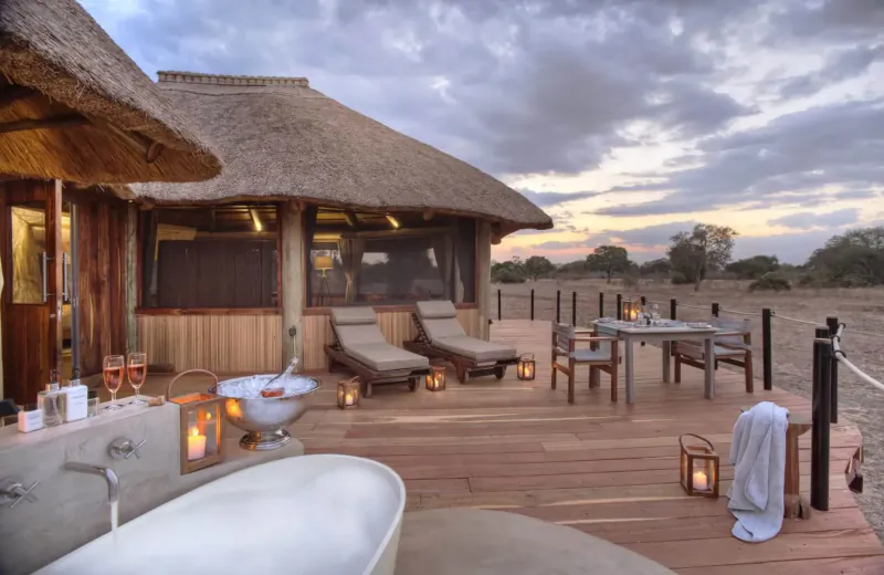 Luxury thatched Lion Camp tent at sunset with outdoor bathtub, loungers, dining table, and lanterns on deck