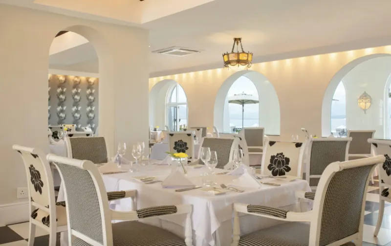 Elegant dining room at The Marine Hotel Hermanus with white tablecloth, floral chairs, chandelier, and ocean-view arches.