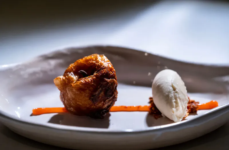Crispy golden pastry dumpling with ice cream scoop, orange sauce drizzle, and crumbs on white plate