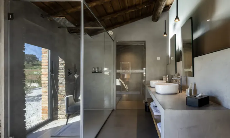 Modern rustic bathroom with glass shower, double white sinks on concrete vanity, wooden beams, and countryside view.