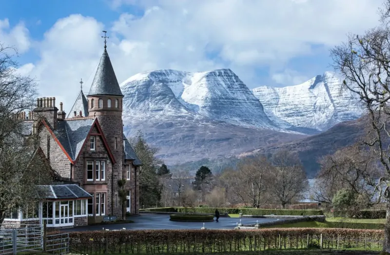 Red sandstone turreted mansion by Loch Lomond with snowy Cuillin mountains under blue sky.