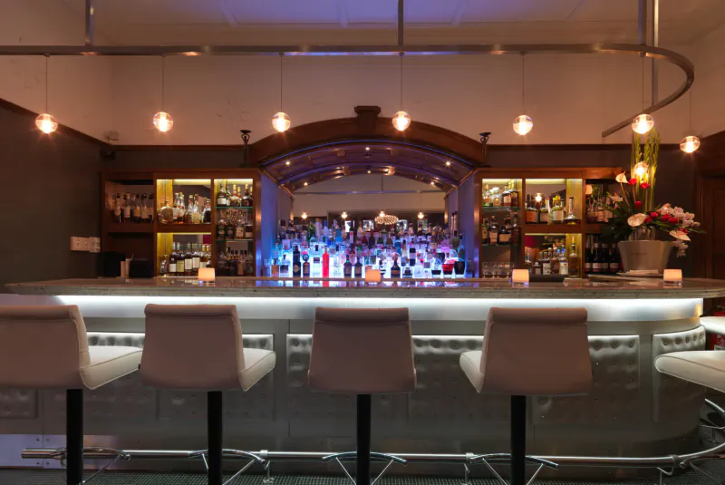 Elegant hotel bar with glowing blue-lit shelves stocked with liquor bottles and white stools at curved counter.