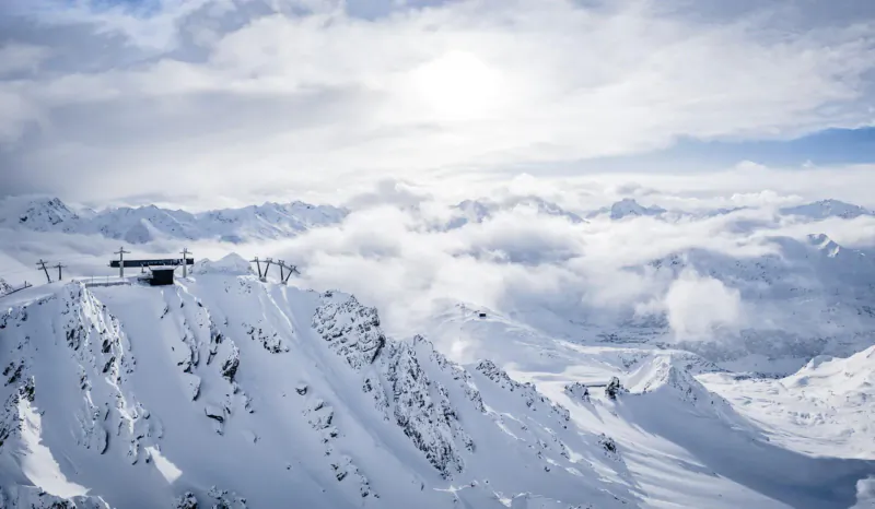 Snowy ski slopes in St Anton am Arlberg with mountain hut, clouds, and distant peaks under partly cloudy sky.