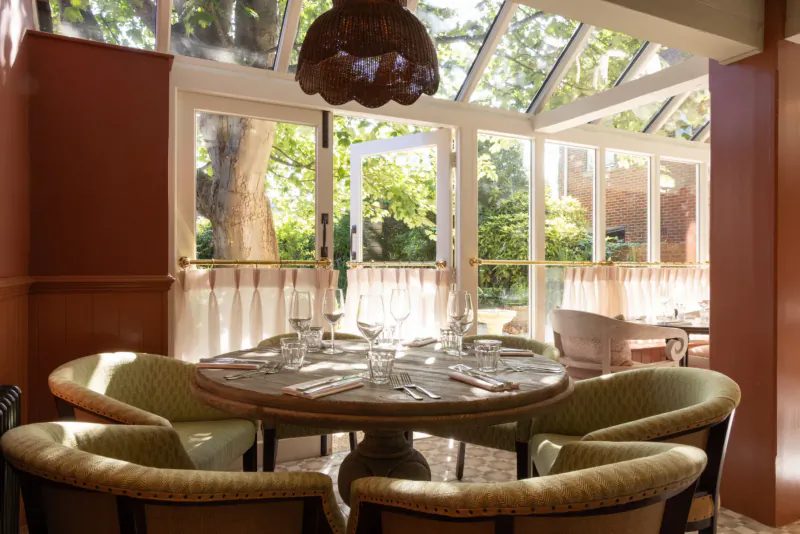 Sunlit conservatory dining room with round wooden table set for four, sheer curtains, garden view at The George & Dragon Marlow.