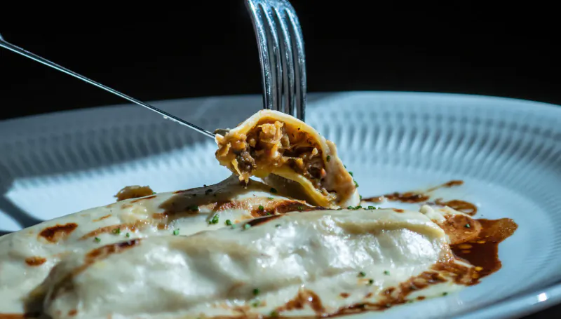 Fork lifting cheesy ravioli with meat filling from white plate garnished with sauce and chives at El Llorenç.