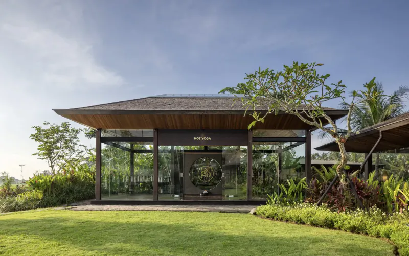 Modern glass pavilion with wooden roof and yin-yang door in lush tropical garden at GDAS Bali retreat, sunny day.