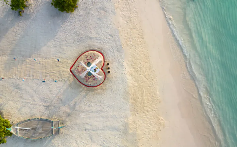 Aerial view of heart-shaped tent with couple inside on white sandy beach by turquoise sea at Amari Raaya Maldives