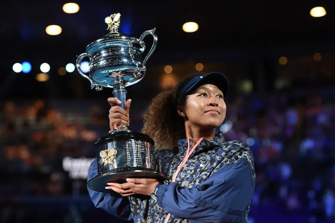Naomi Osaka smiles triumphantly holding the Australian Open trophy on court, wearing cap and jacket.